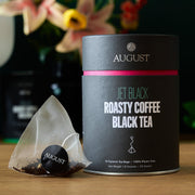 August Jet Black Roasty Coffee Black Tea packaging with a tea bag on a wooden surface.