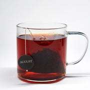 Clear glass mug with red tea and an August tea bag on a white background