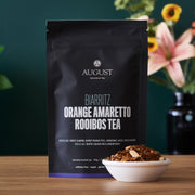 August Biarritz Orange Amaretto Rooibos Tea package with a bowl of tea leaves on a wooden surface.