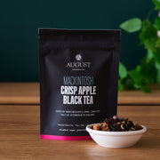 August Mackintosh Crisp Apple Black Tea package with a bowl of tea leaves on a wooden surface.