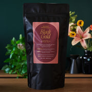 Black tea package labeled 'Black Gold' by August Essentials on a wooden surface with plants in the background.
