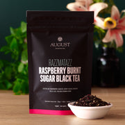 August tea package for Razzmatazz Raspberry Burnt Sugar Black Tea on a wooden surface with tea leaves.