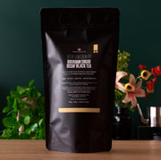 Black tea package with text on a wooden surface and blurred greenery in the background