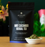 August Olympus Mint Cucumber Herbal Tea package with tea leaves in a bowl on a wooden surface.