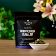 August Olympus Mint Cucumber Herbal Tea package with tea leaves in a bowl on a wooden surface.