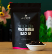 August Southern Gothic Peach Bourbon Black Tea package with a small bowl of tea leaves on a wooden surface.