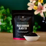 August Southern Gothic Peach Bourbon Black Tea package with tea leaves in a bowl on a wooden surface.