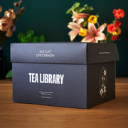 Tea library box with 'August Uncommon' branding on a wooden surface with flowers in the background