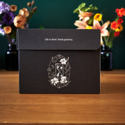 Black box with tea-themed design and quote on a wooden surface with flowers in the background