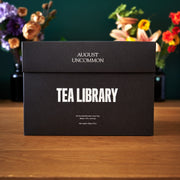Black box labeled 'August Uncommon Tea Library' on a wooden surface with flowers in the background.