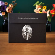 Black box with white emblem and text on a wooden surface with flowers in the background