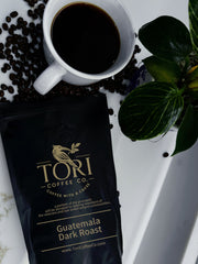 Tori Coffee Co. Guatemala Dark Roast coffee bag with a cup of coffee and coffee beans on a white surface.