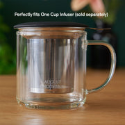 Clear glass mug with metal infuser on a wooden surface, with text about compatibility.