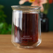 Glass jar with dark liquid labeled 'August Uncommon' on a wooden surface with a blurred background.