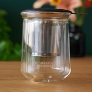 Clear glass tea infuser with metal mesh on a wooden surface, branded 'August Uncommon'.