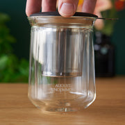 Clear glass tea infuser with metal mesh and black lid on a wooden surface, branded 'August Uncommon'.