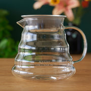 Clear glass coffee pitcher on a wooden surface with a blurred floral background