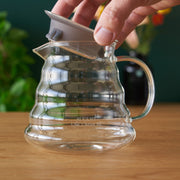 Clear glass pitcher with handle on a wooden surface, hand holding a white lid.