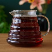 Glass pitcher filled with a dark liquid on a wooden surface with a blurred floral background