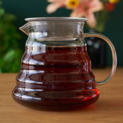 Clear glass coffee carafe with a handle filled with coffee on a wooden surface, blurred flowers in the background.