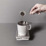 Person adding tea leaves to a white mug on a scale with a gray background