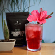 Red drink in a glass with a pink flower, August Smoky Hibiscus Cherry Tea package, and cactus plant on a wooden surface.