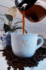 Coffee being poured into a white mug with 'TORI Coffee Co.' logo, surrounded by coffee beans and plants.