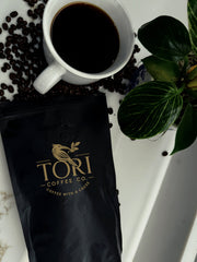 White mug of coffee with Tori Coffee Co. bag and scattered coffee beans on a white surface.