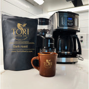 Tori Coffee Co. dark roast coffee package, mug, and coffee maker on a kitchen counter.