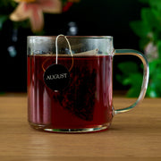 Clear mug with red liquid and a tea bag labeled 'August' on a wooden surface.