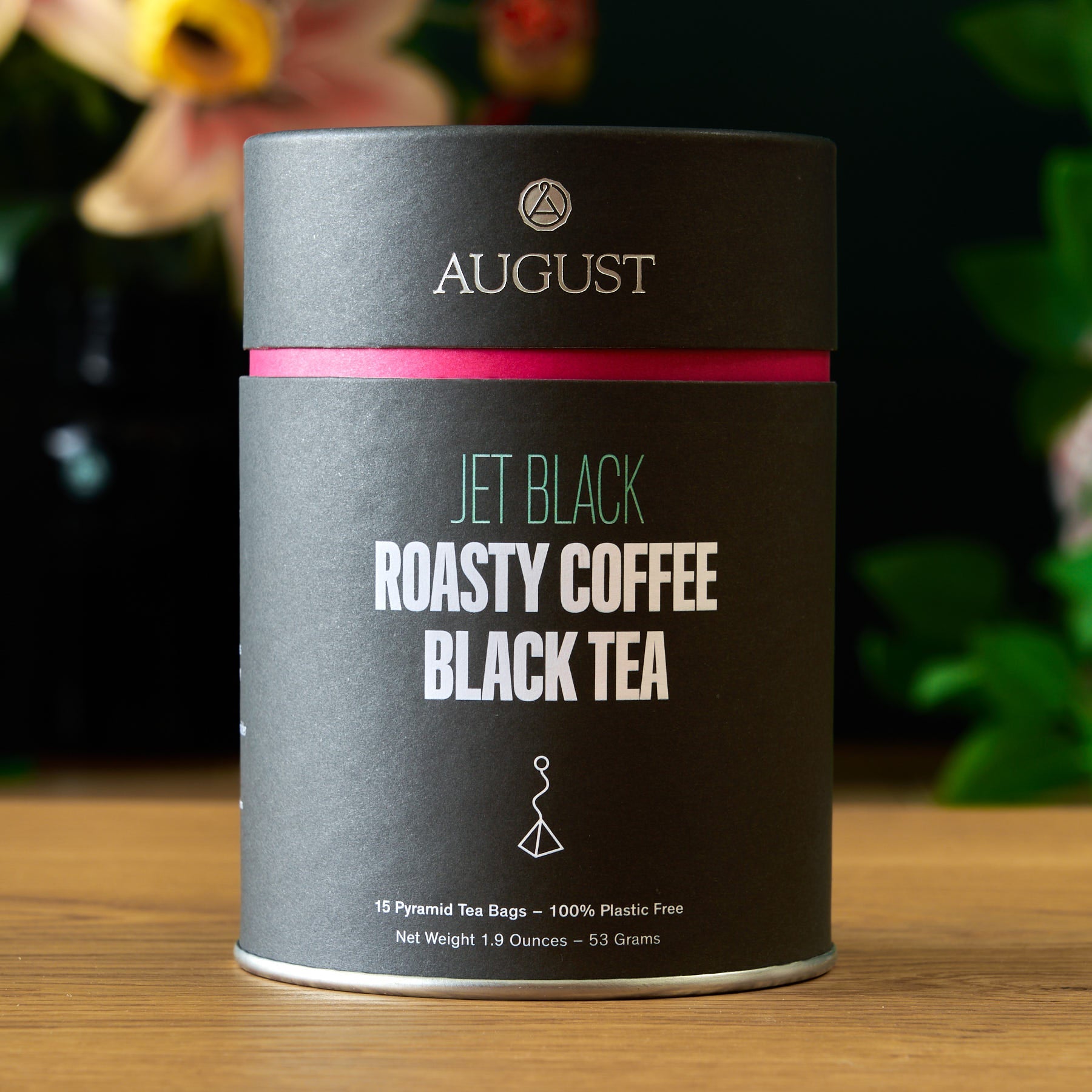 August Jet Black Roasty Coffee Black Tea canister on a wooden surface with a blurred floral background