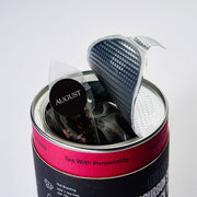Tea bag with a strainer in a can labeled 'August Tea' on a light gray background