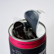 Tea bag with a strainer in a can labeled 'August Tea' on a light gray background