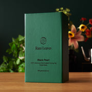 Green tea box labeled 'Rare Leaves Black Pearl' on a wooden surface with a blurred floral background