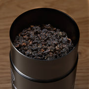 Black container filled with dark tea leaves on a wooden surface