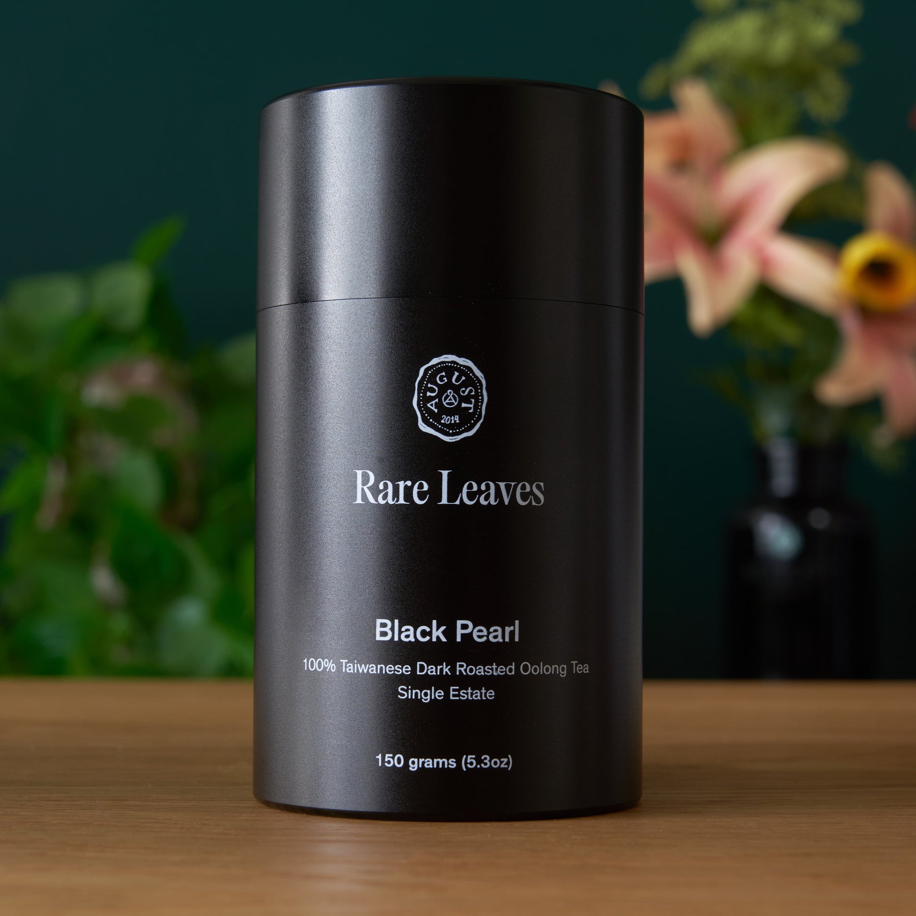 Black tea canister labeled 'Rare Leaves Black Pearl' on a wooden surface with a blurred floral background.