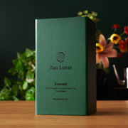 Green tea box labeled 'Rare Leaves Emerald' on a wooden surface with a blurred floral background