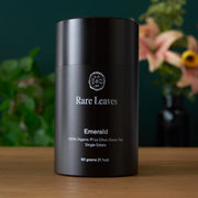 Black container labeled 'Rare Leaves Emerald' on a wooden surface with a blurred floral background