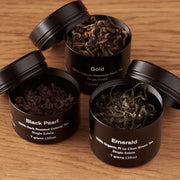 Four black tea tins labeled 'Gold', 'Black Pearl', and 'Emerald' on a wooden surface.