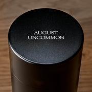 Black cylindrical container with 'August Uncommon' text on a wooden surface