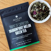 August Tea package for 'Tropic of Capricorn Dragonfruit Melon Green Tea' with a bowl of tea leaves on a wooden surface.